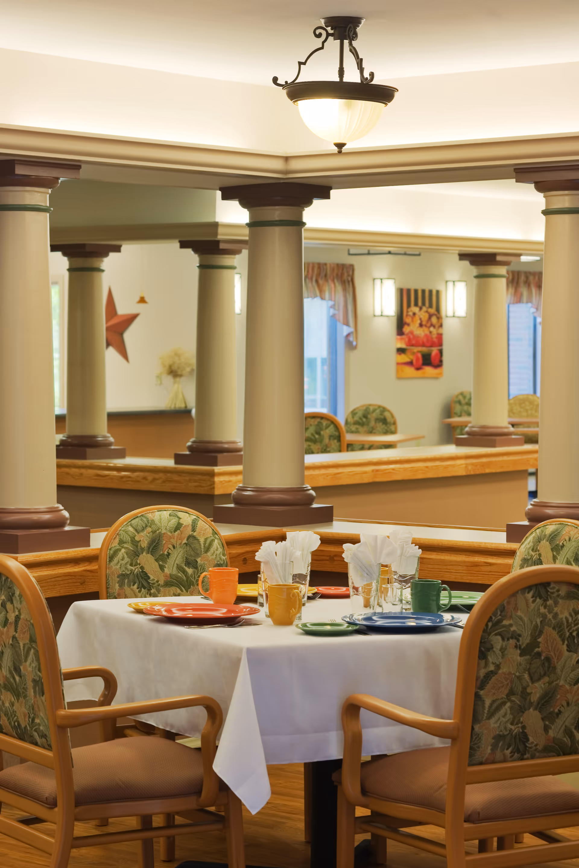 A dining area in Oak Grove Center with a table set for four. The table has a white tablecloth, colorful plates, mugs, and napkins. The chairs have wooden frames with floral patterned upholstery. The room features decorative columns, soft lighting, and wall art, creating a warm and inviting atmosphere.