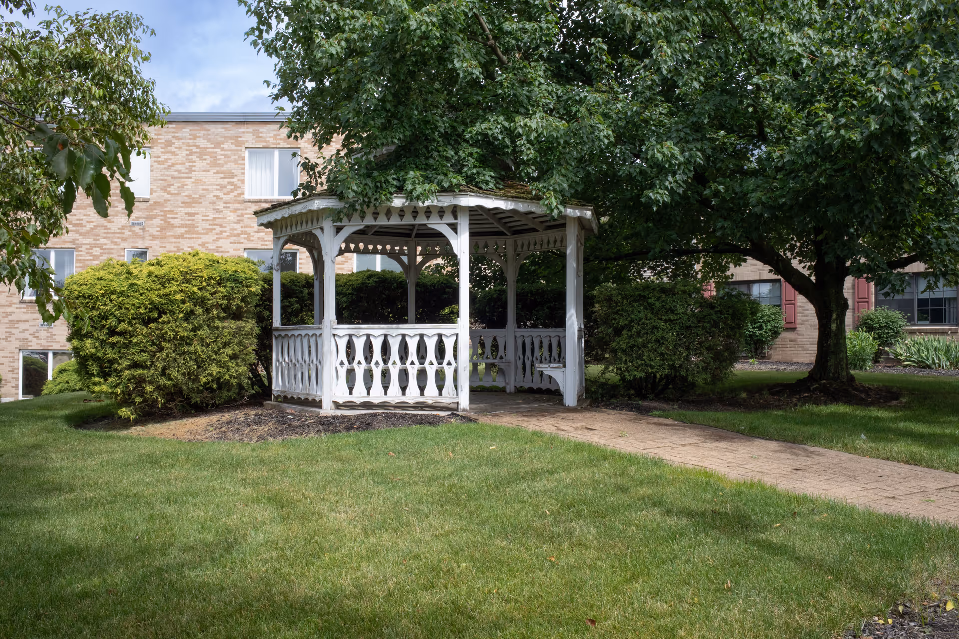 A white wooden gazebo with decorative railings situated on a grassy lawn surrounded by green bushes and trees. A paved walkway leads up to the gazebo. In the background, there is a brick building with several windows.