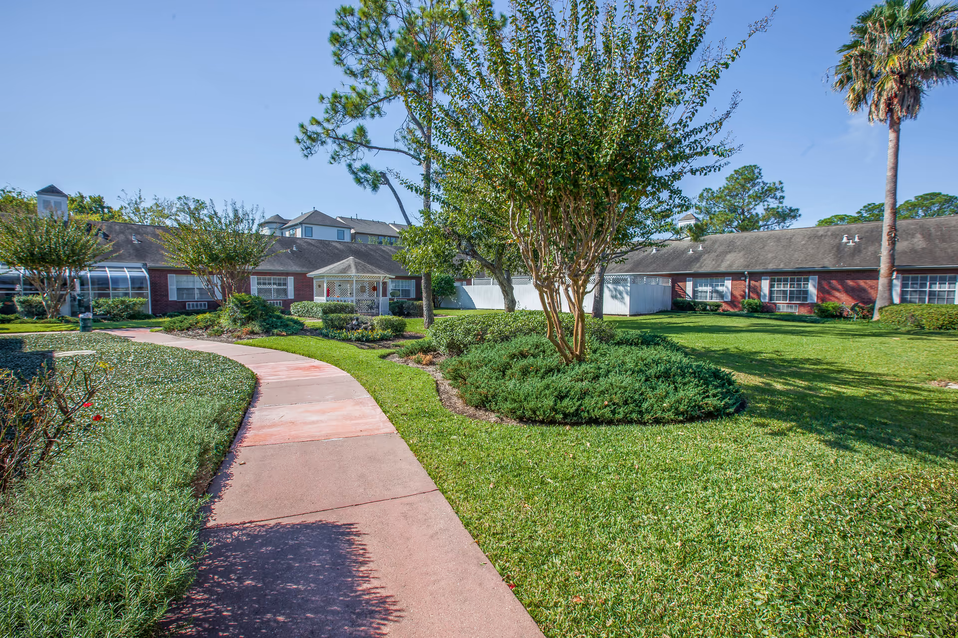 A landscaped outdoor area at Brookdale Memorial Oaks featuring a curved concrete pathway, green grass, bushes, and several trees including a palm tree. In the background, there are single-story brick buildings with multiple windows and a small white gazebo.