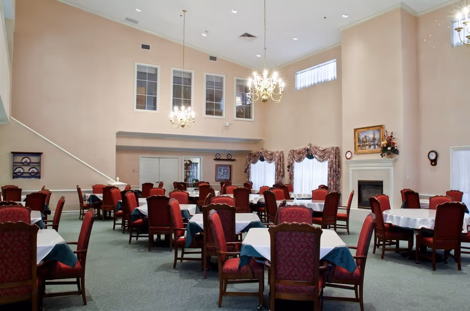A spacious dining room with multiple tables covered in white and green tablecloths, surrounded by red upholstered chairs. The room features high ceilings with two chandeliers, large windows with floral curtains, a fireplace with a painting and floral arrangement above it, and a clock on the wall.
