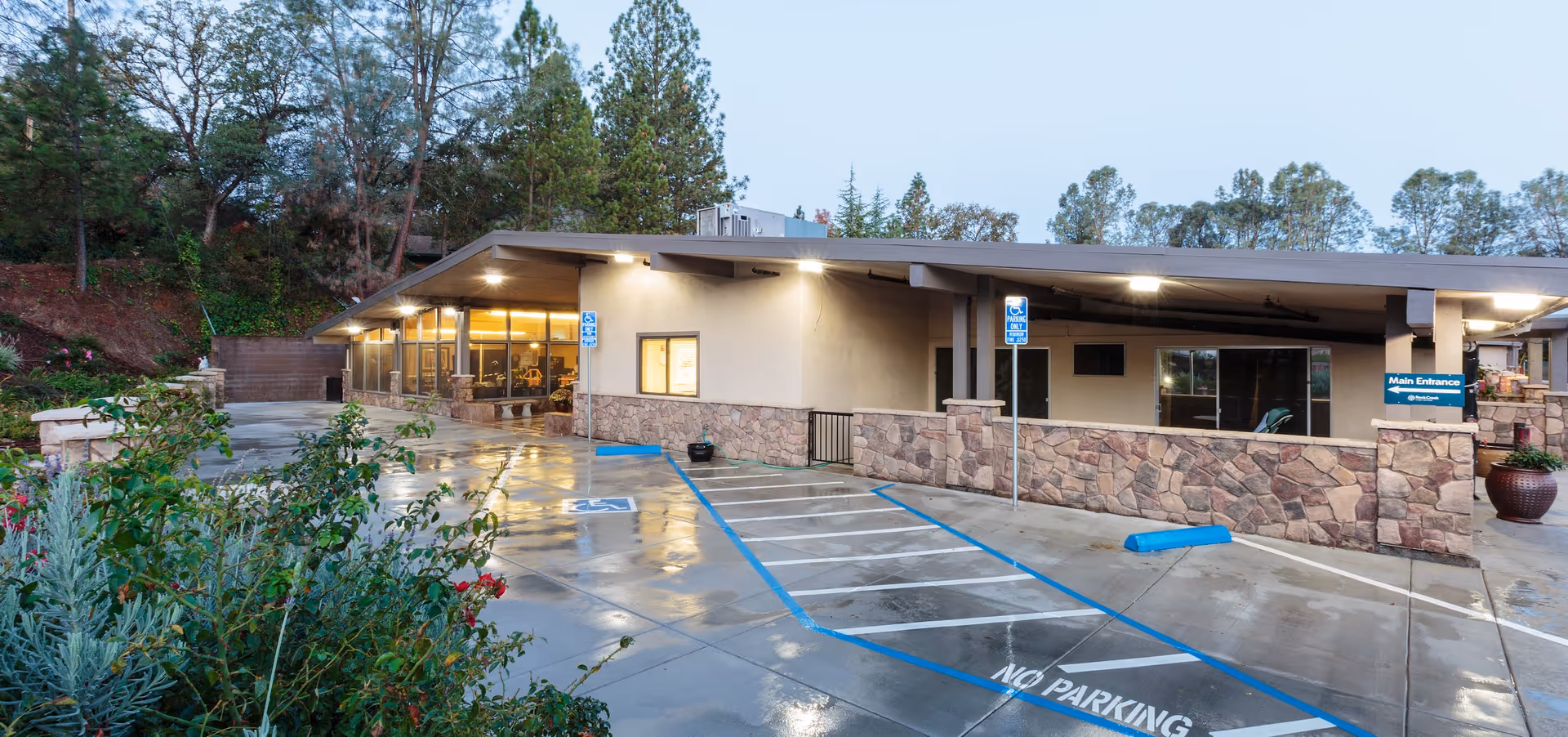 Exterior view of Rock Creek Care Center building during early evening with wet pavement, accessible parking spaces, stone facade, and surrounding trees.