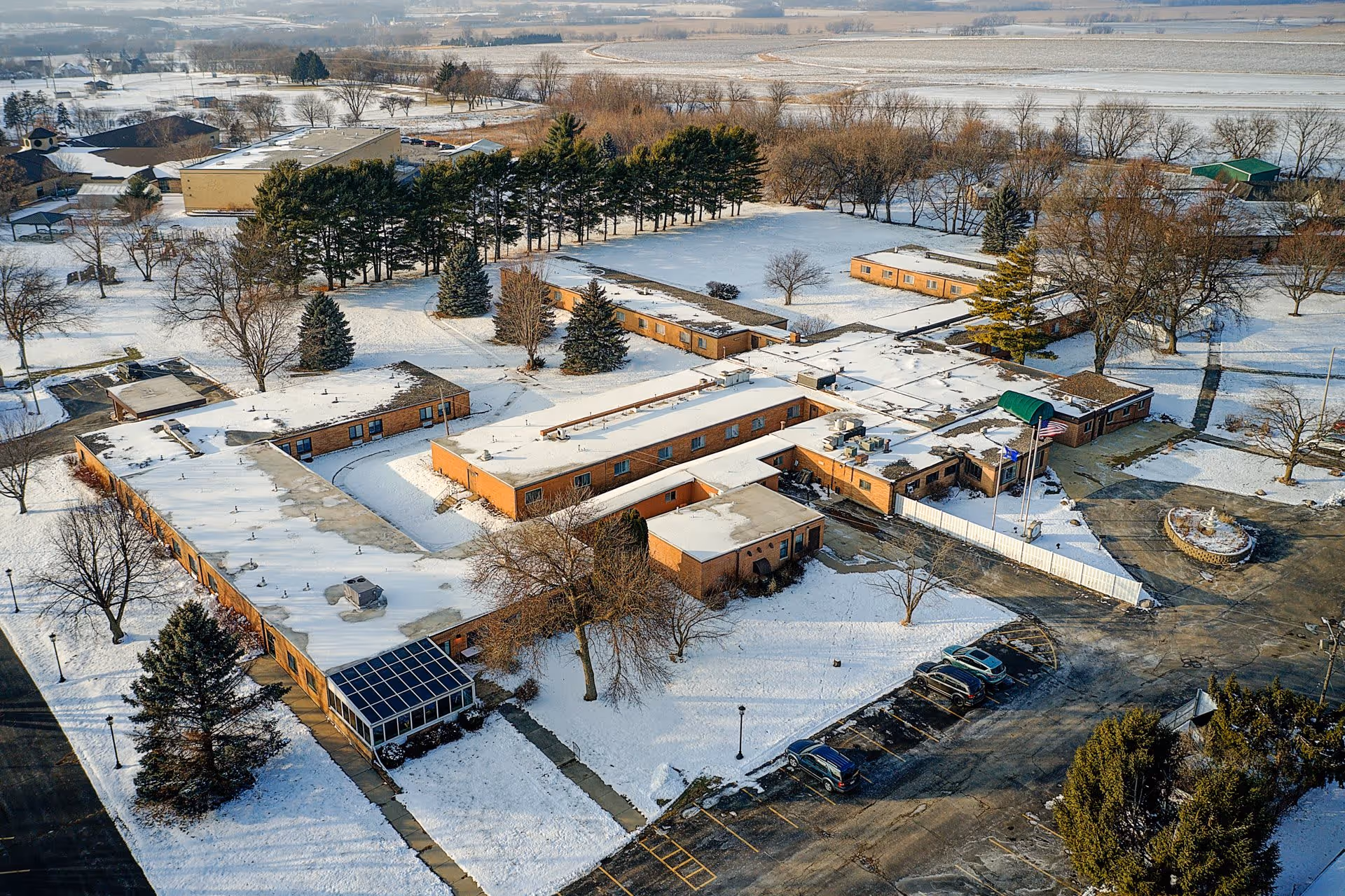 Aerial view of Wolverton Glen senior living facility covered in snow, showing multiple connected single-story brick buildings with flat roofs, surrounded by trees and parking areas with several cars parked.