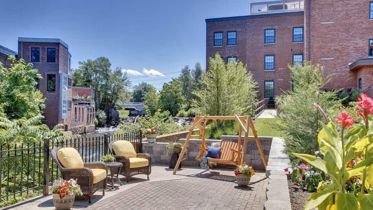 Outdoor patio area at Atria Marland Place featuring two wicker chairs with yellow cushions, a small round table, a wooden swing with cushions, potted plants, and greenery surrounding the space with brick buildings in the background under a clear blue sky.