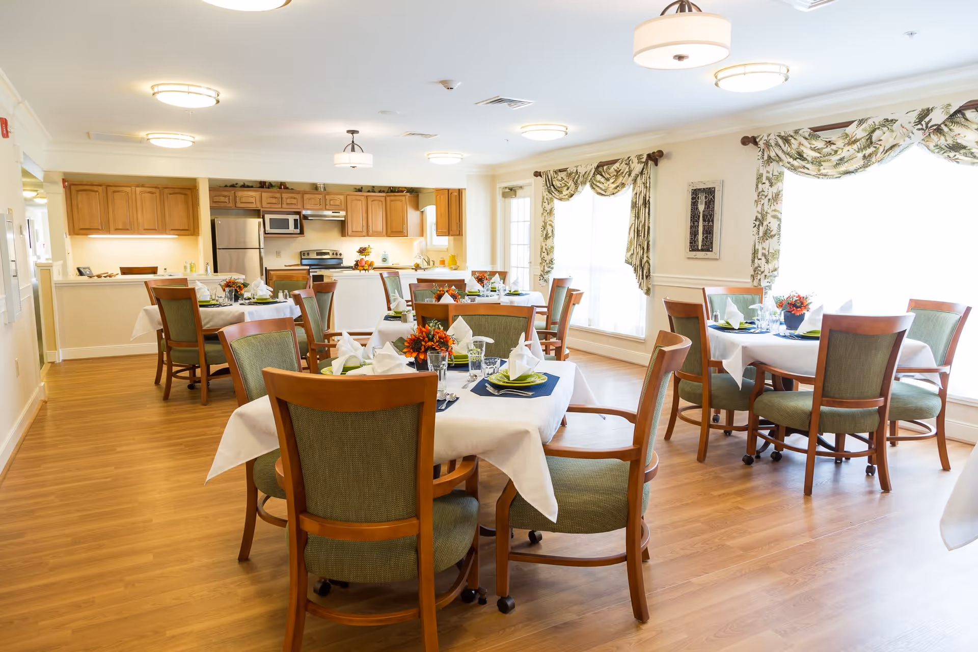 Bright dining room with several round tables covered with white tablecloths and set with plates, napkins, glasses, and floral centerpieces. The room has wooden floors, green cushioned wooden chairs, large windows with floral curtains, and a kitchen area with wooden cabinets and stainless steel appliances in the background.