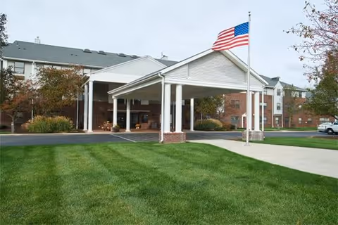 Exterior view of a multi-story senior living facility with a covered entrance, an American flag on a flagpole, well-maintained green lawn, and trees with autumn foliage.
