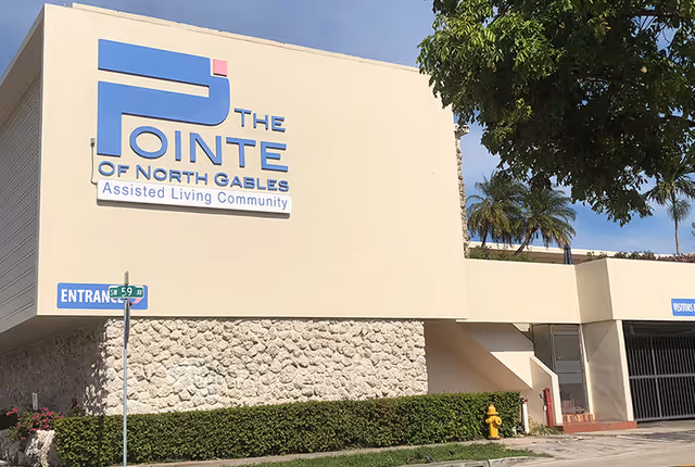 Exterior view of The Pointe of North Gables Assisted Living Community building with a large sign on the wall, a street sign indicating entrance, green bushes, a fire hydrant, and palm trees in the background under a clear sky.