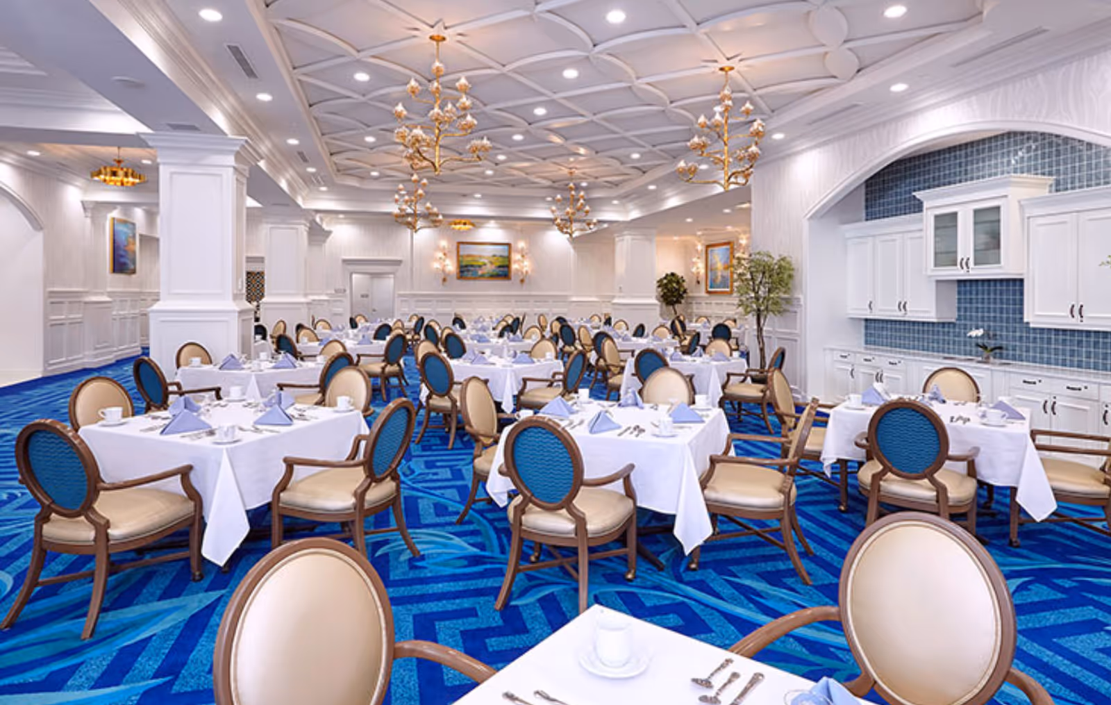 A spacious, elegant dining room with multiple tables covered in white tablecloths, each set with blue napkins, cups, and silverware. The chairs have beige seats and blue backs. The room features a vibrant blue patterned carpet, white paneled walls, decorative ceiling with recessed lighting, and gold chandeliers. There are paintings and plants decorating the walls and corners.