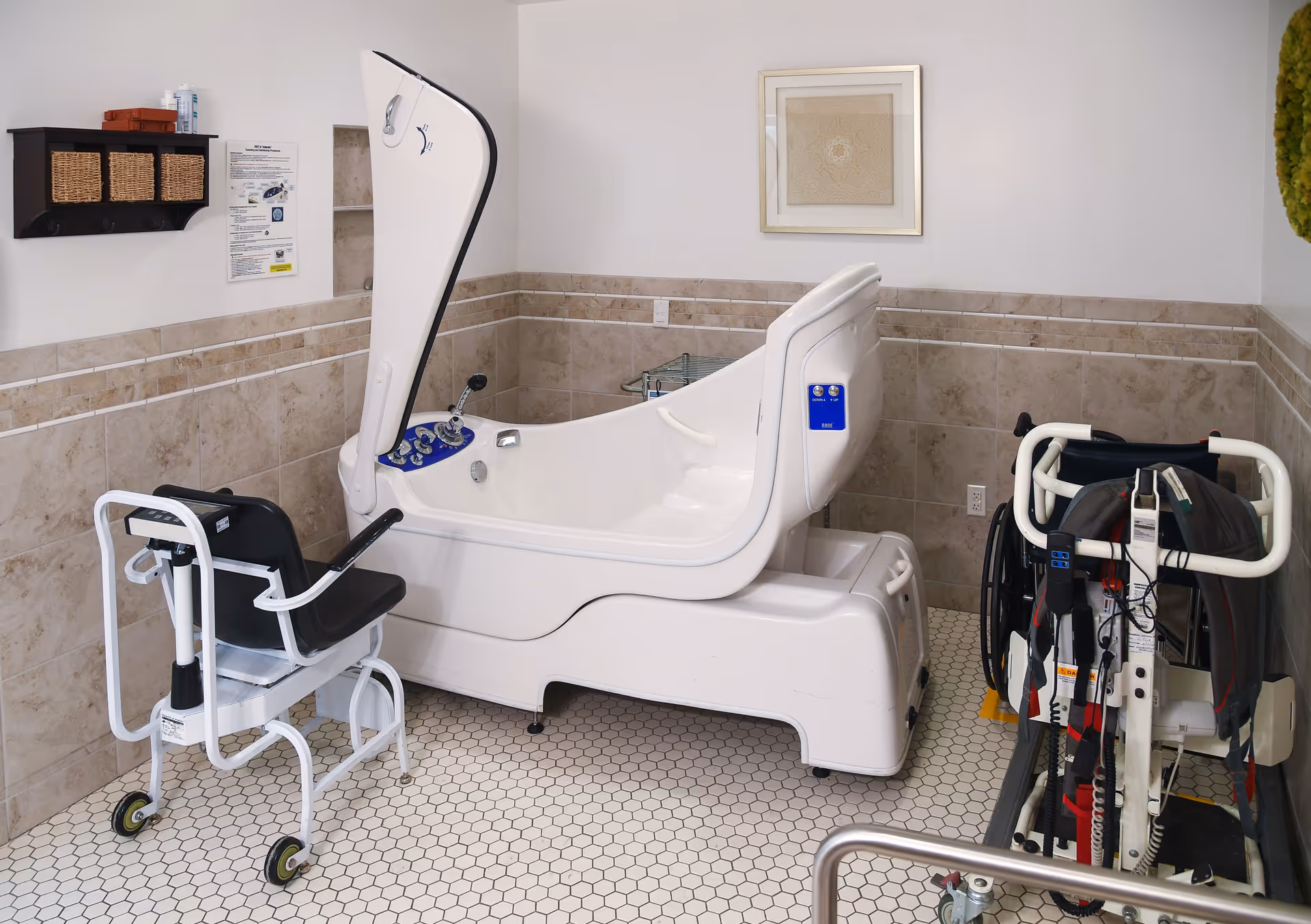 A specialized bathing room in a senior living facility featuring a large white assisted bathing tub with a raised lid, a black and white shower chair with wheels, and a wheelchair with support straps. The room has beige tiled walls, white hexagonal floor tiles, a small wall shelf with baskets, and a framed artwork on the wall.