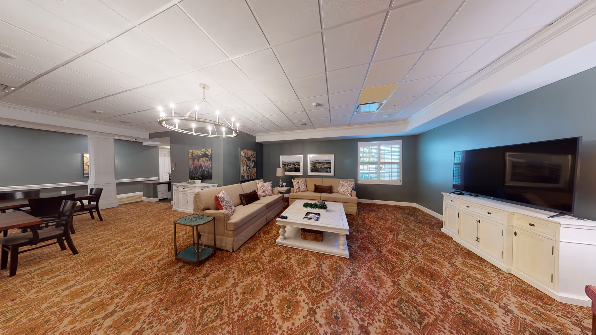 A spacious senior living common area with two beige sofas arranged around a white coffee table on a patterned carpet. The room features a large flat-screen TV on a white cabinet, a circular chandelier, framed artwork on the walls, and a window with white shutters letting in natural light. There are also dining tables and chairs visible on the left side of the room.