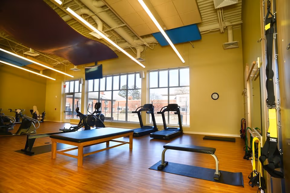A spacious fitness room with large windows letting in natural light. The room features exercise equipment including treadmills, rowing machines, a workout bench, and resistance bands hanging on the wall. The ceiling has exposed pipes and modern lighting fixtures.