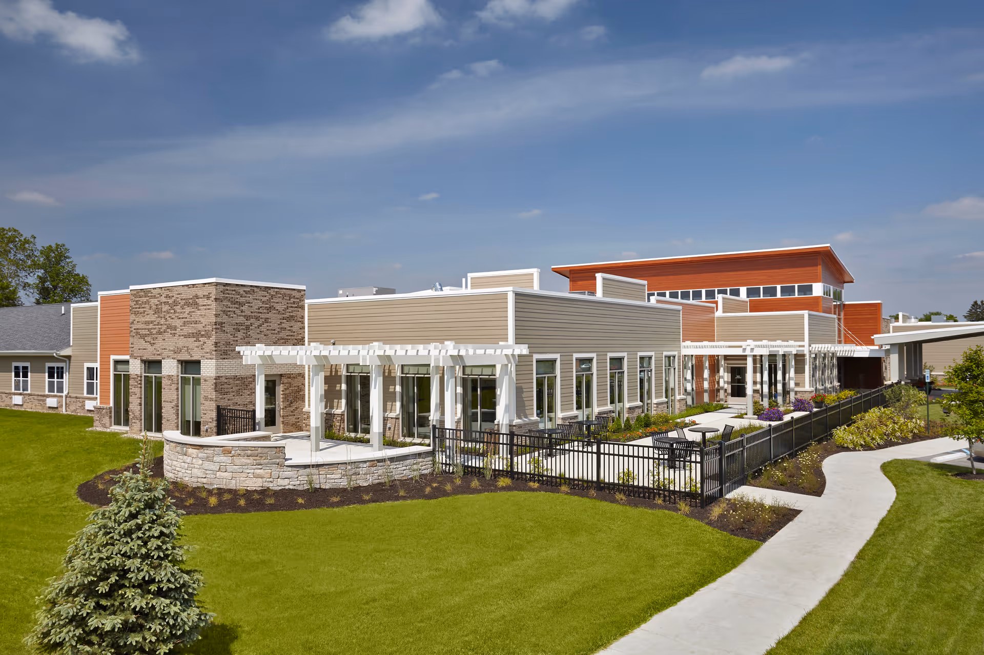 Exterior view of The Springs at Lafayette senior living facility showing a modern building with beige and brick walls, large windows, a white pergola, outdoor seating area, well-maintained green lawn, and a curved concrete walkway under a partly cloudy blue sky.