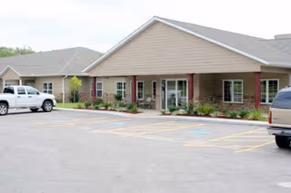 Exterior view of a single-story senior living facility building with a covered entrance, beige siding, and several windows. There are two vehicles parked in the parking lot in front of the building, and some landscaping with small bushes and plants near the entrance.