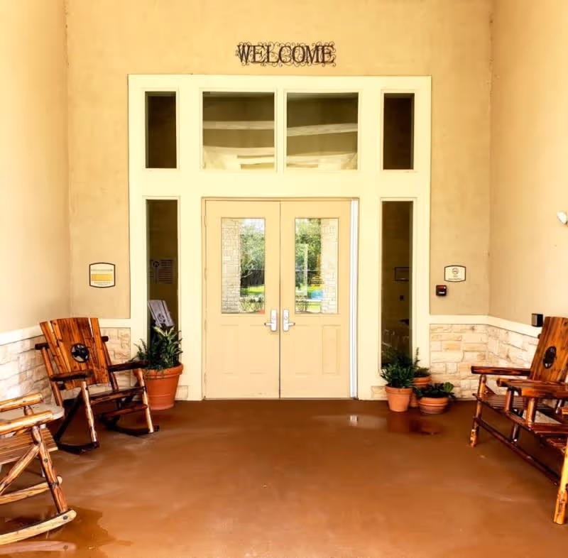 Entrance area with double doors and a 'WELCOME' sign above. There are wooden rocking chairs and potted plants on either side of the doors, with beige walls and stone accents.