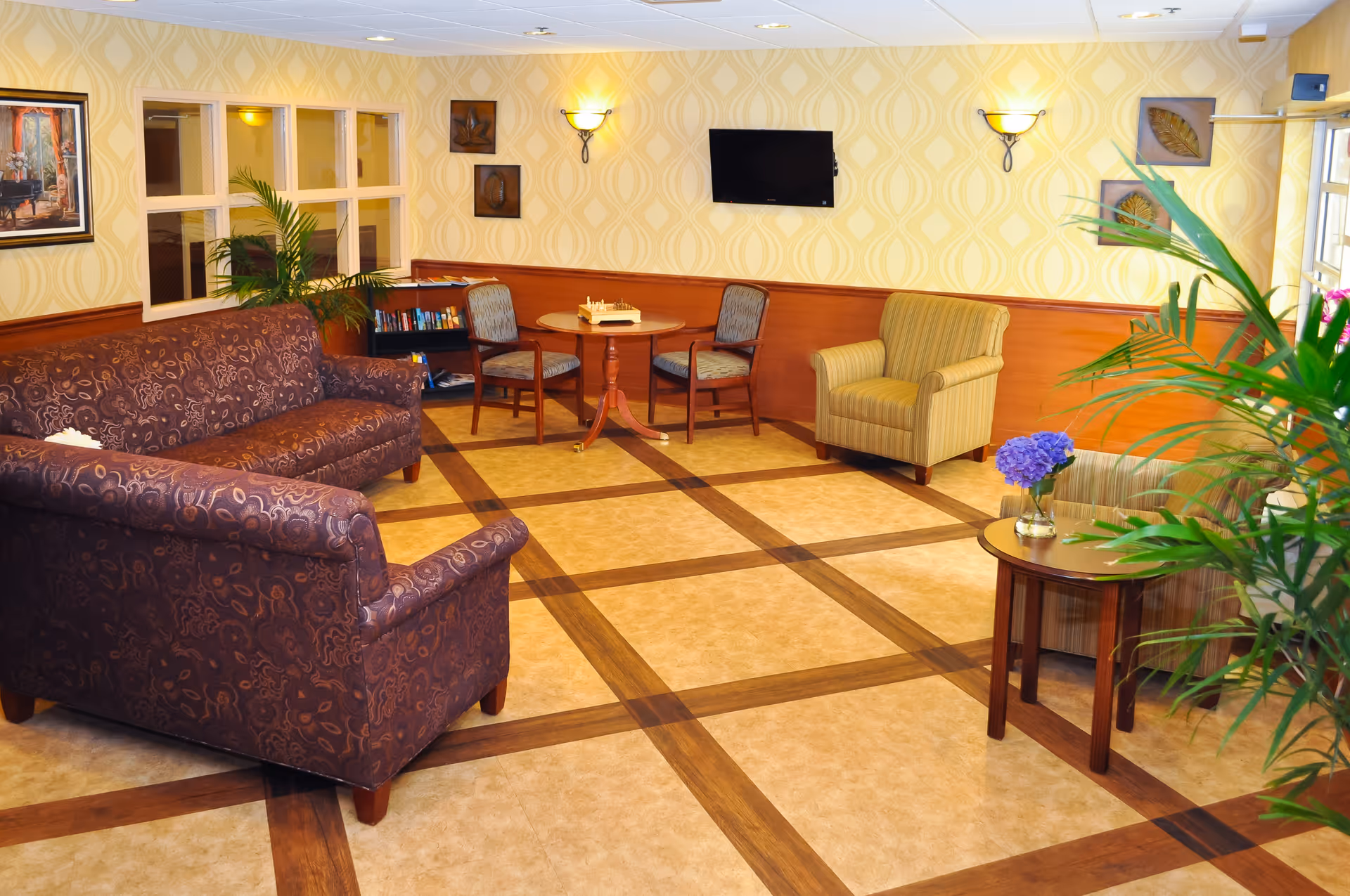 A cozy living room area with patterned wallpaper and wood paneling. The room features a brown patterned sofa, a striped armchair, two wooden chairs around a small round table with a chessboard, a small side table with a vase of purple flowers, a flat-screen TV mounted on the wall, and several green plants. The floor has a beige and brown checkered pattern.