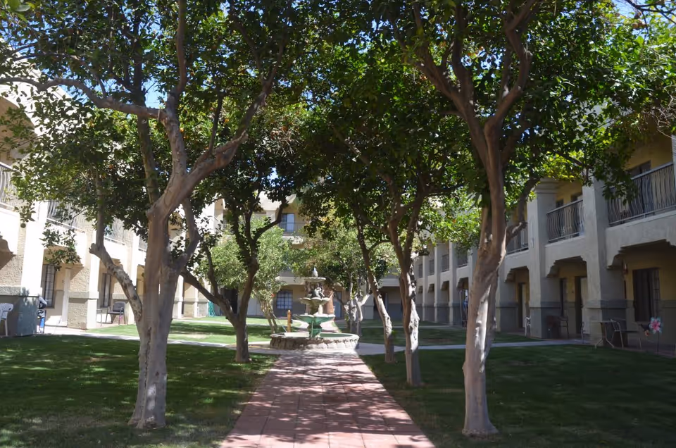 A courtyard area in an assisted living facility with a tiled walkway lined by trees on both sides leading to a multi-tiered water fountain. The courtyard is surrounded by two-story buildings with balconies and doors opening to the outdoor space. The grass is well-maintained and the area is shaded by the trees.