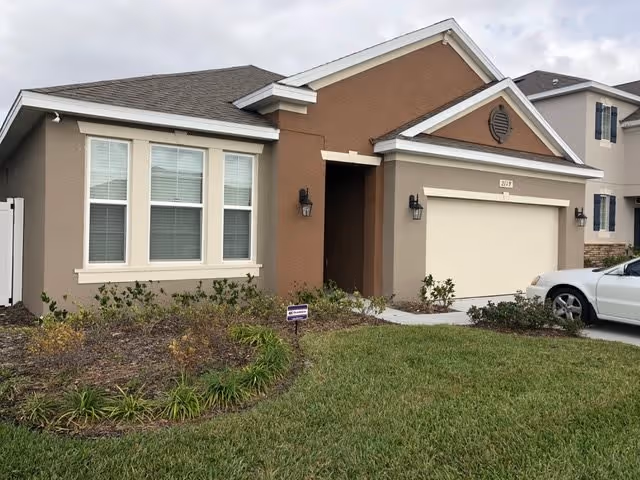 Front exterior view of a single-story house with beige and brown walls, a two-car garage, and a well-maintained lawn with shrubs and plants. A white car is parked in the driveway.