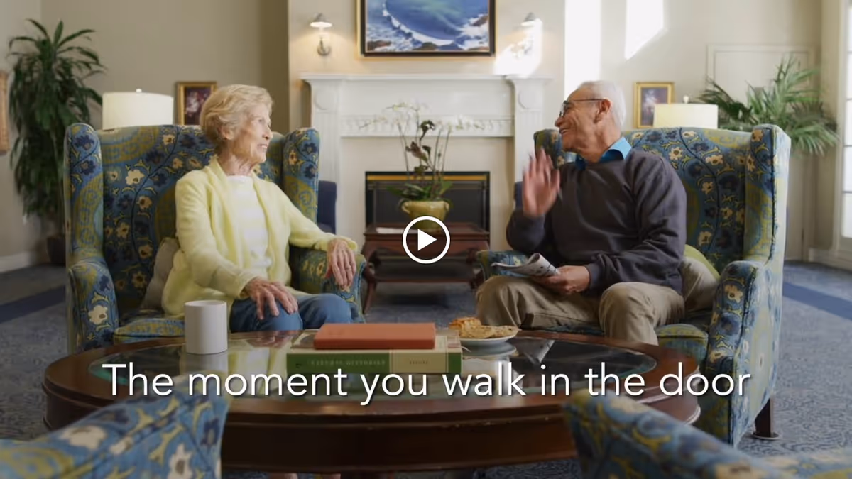 Two elderly people sitting in patterned armchairs facing each other in a cozy living room with a fireplace, plants, and framed pictures on the walls. A coffee table with books, a cup, and a plate of cookies is in front of them. Text on the image reads 'The moment you walk in the door.'
