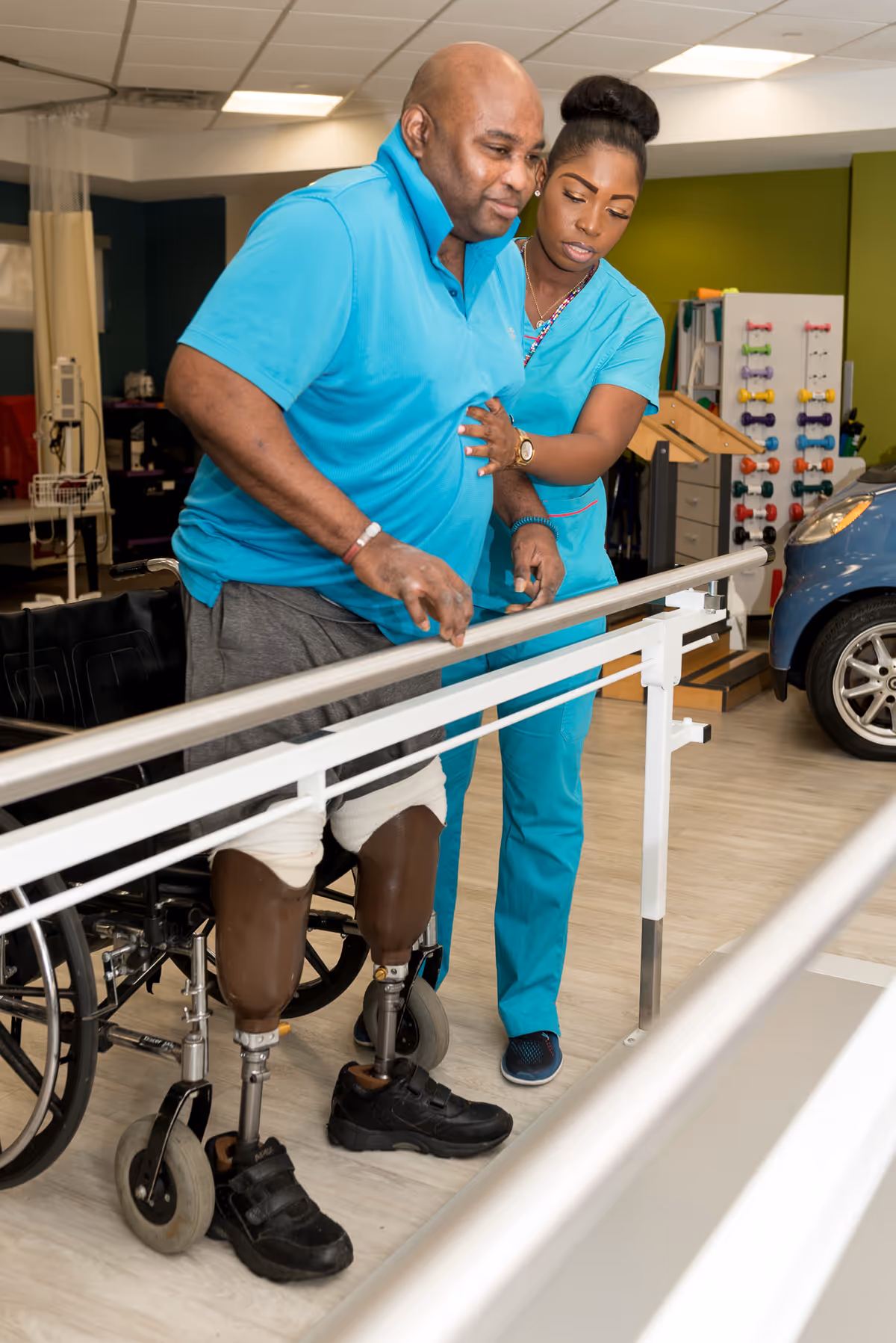 A man with double leg prosthetics practices walking with parallel bars while being assisted by a female healthcare worker in a rehabilitation center. A wheelchair is visible behind him and various therapy equipment is in the background.