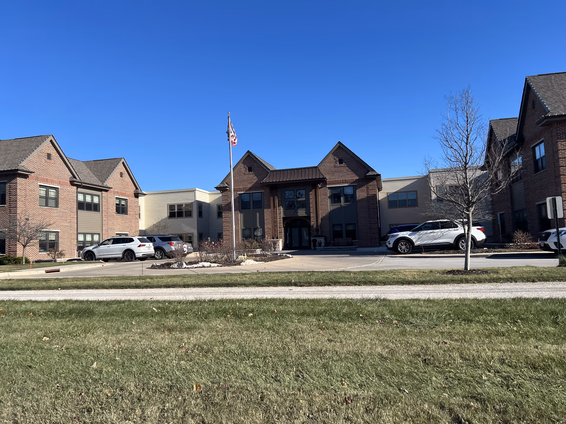 Exterior view of a senior living facility building with brick and beige siding, several parked cars, a flagpole with an American flag, and a clear blue sky above.