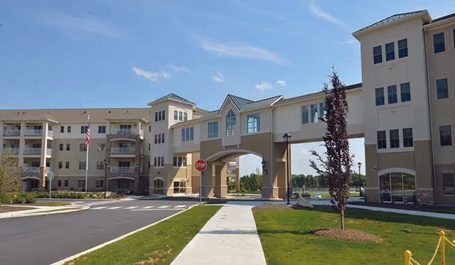 Exterior view of The Long Community at Highland showing a multi-story building with a covered archway connecting two sections. There is a sidewalk, a stop sign, and landscaped grass areas with small trees under a clear blue sky.