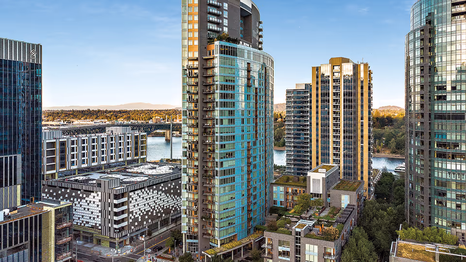 View of a cityscape featuring several modern high-rise buildings with glass facades, a river in the background, and a bridge spanning the river. The scene is taken during daylight with clear skies.