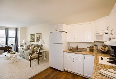 A bright and clean apartment interior featuring a small kitchen area with white cabinets, a white refrigerator, microwave, and stove. The kitchen has a granite countertop with various kitchen items. Adjacent to the kitchen is a living room area with a beige sofa, two framed pictures on the wall, a white coffee table, a wooden chair, and a window with blue curtains letting in natural light.