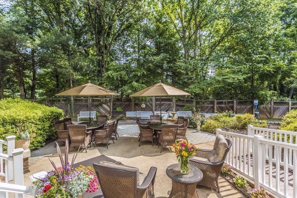 Outdoor patio area with multiple wicker chairs and tables, two large beige umbrellas, surrounded by lush green trees and bushes. There are white benches along a wooden fence in the background and colorful flowers in planters near the seating area.
