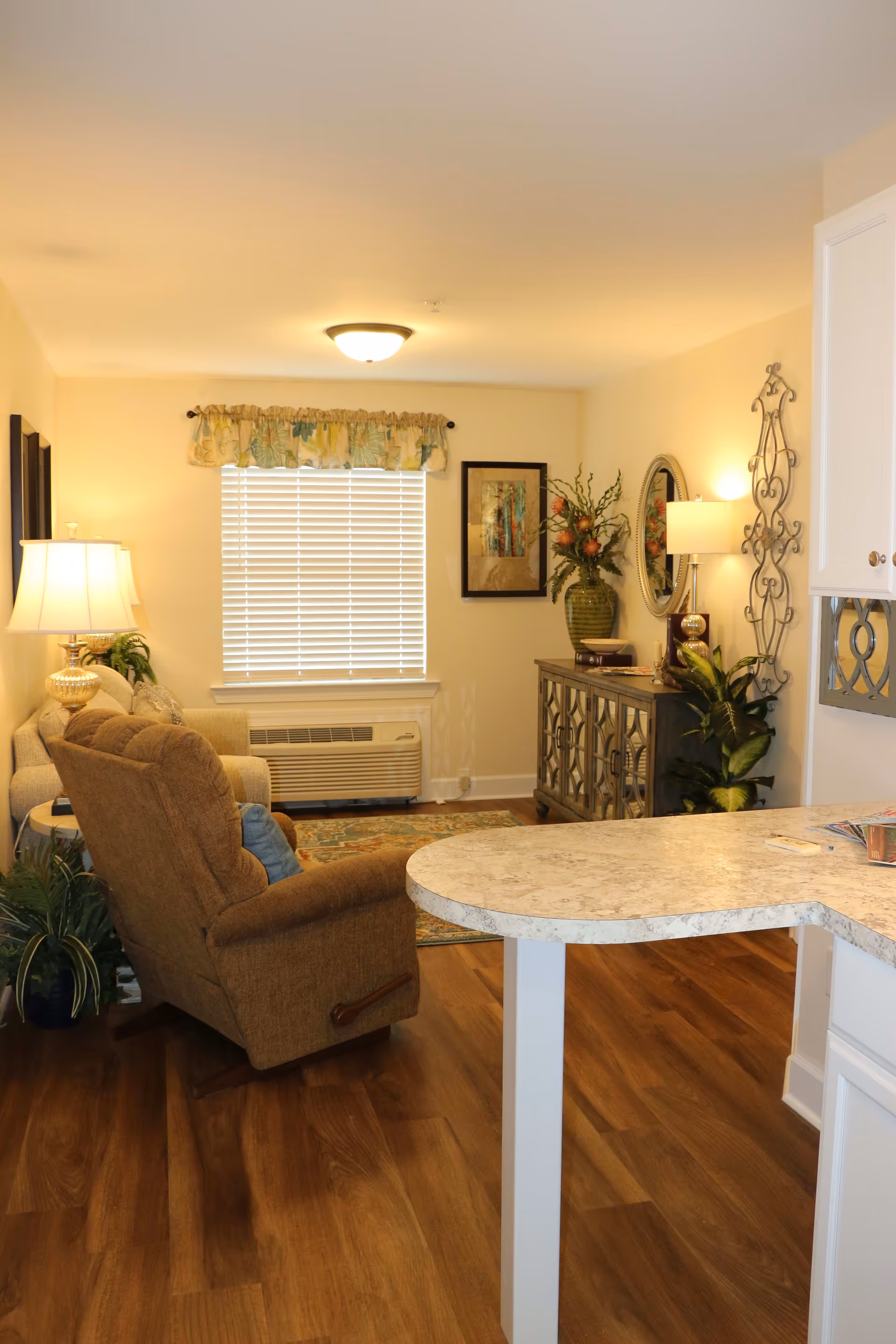 A cozy living room area with a brown recliner chair, a beige sofa, a side table with a lamp, and a window with blinds and a floral valance. There is a decorative cabinet with a large vase of flowers and a round mirror on the wall. The room has wooden flooring and warm lighting.