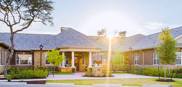 Front exterior view of Silverado Barton Springs Memory Care Community building with a landscaped entrance, trees, benches, and a pathway leading to the main door during daylight.
