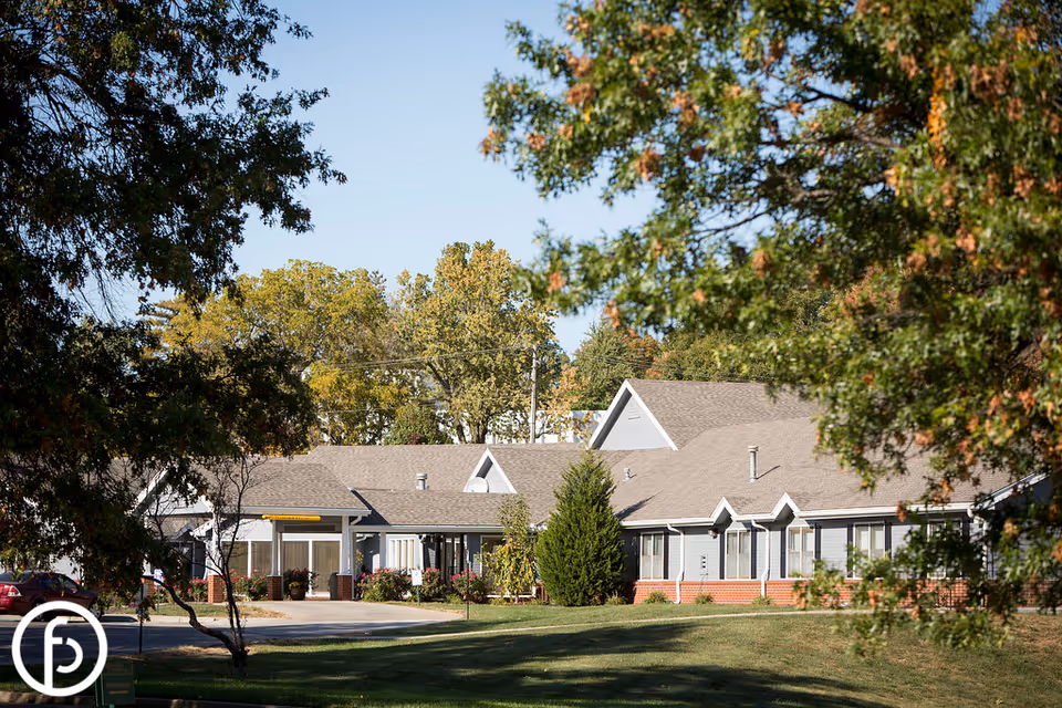Exterior view of a single-story senior living facility building with a gray roof and light-colored walls, surrounded by green trees and a well-maintained lawn under a clear blue sky.
