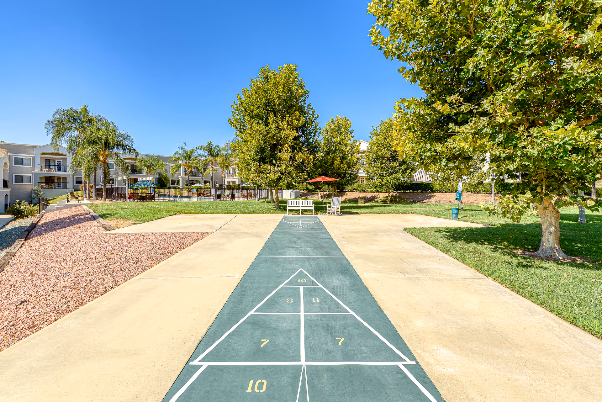 Outdoor shuffleboard court with numbered sections, surrounded by green grass, trees, and benches with umbrellas. In the background, there are multi-story residential buildings with balconies and palm trees under a clear blue sky.
