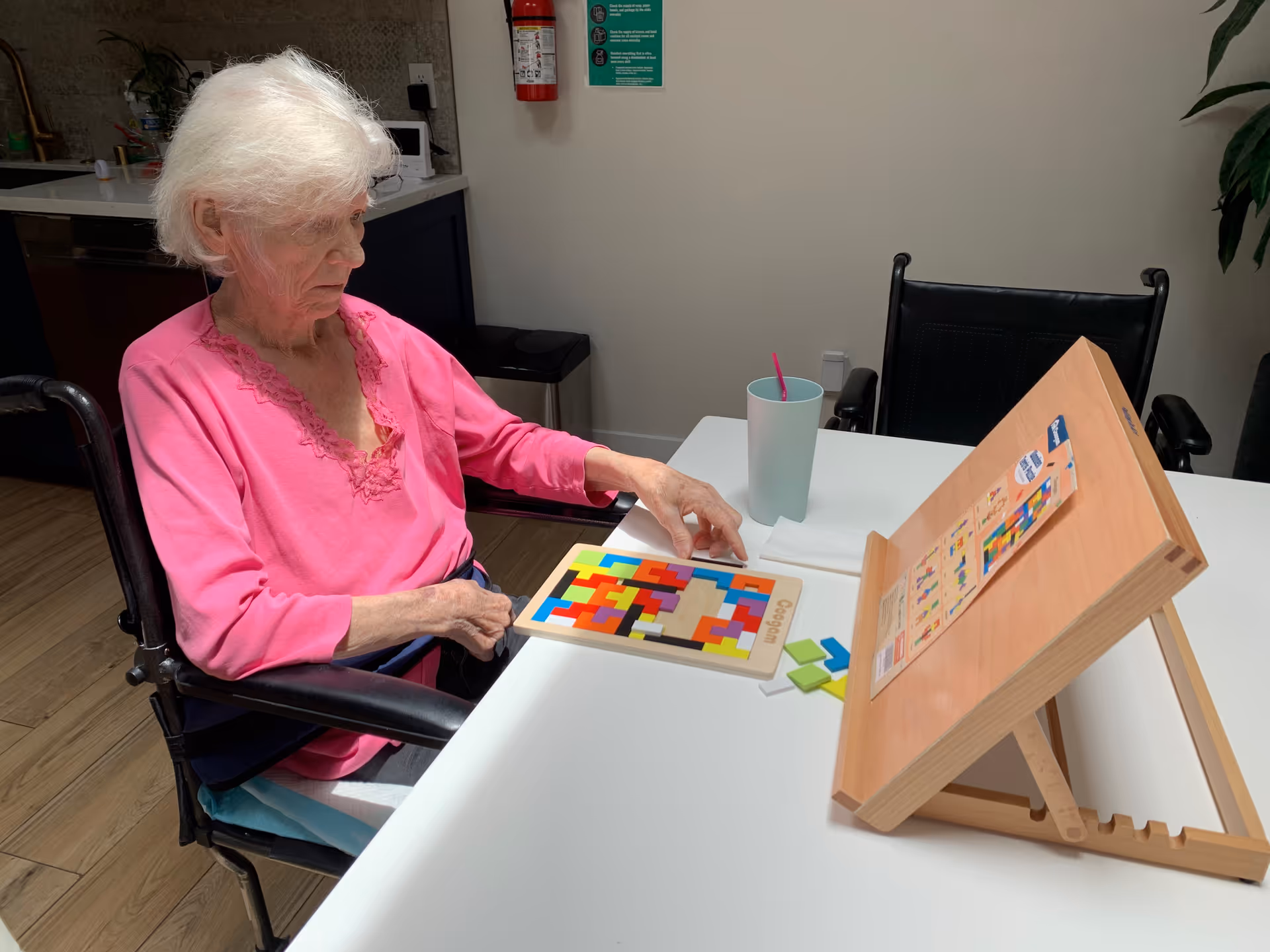 An elderly woman with white hair wearing a pink top is sitting in a wheelchair at a white table, engaging with a colorful wooden puzzle. On the table, there is a light blue cup with a pink straw and a wooden stand holding a booklet or instructions. The background shows part of a kitchen area and a beige wall with a green informational poster.