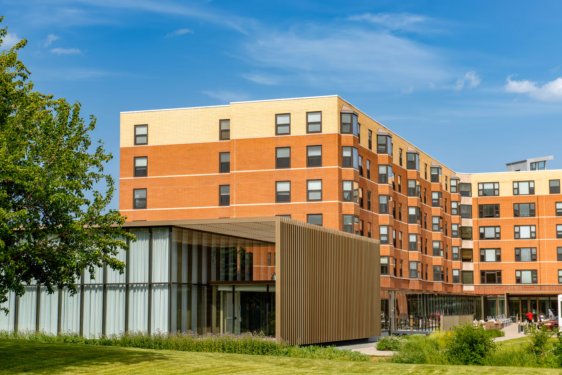 Exterior view of a multi-story retirement center building with a modern glass and wood entrance, surrounded by green grass and trees under a blue sky.