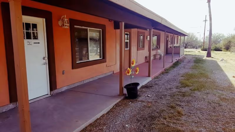 Exterior view of a single-story building with an orange and brown facade, a covered walkway supported by wooden posts, several windows, and a white door with a small sign. There is a potted plant with artificial sunflowers on the concrete walkway and a gravel and grass area beside the building.