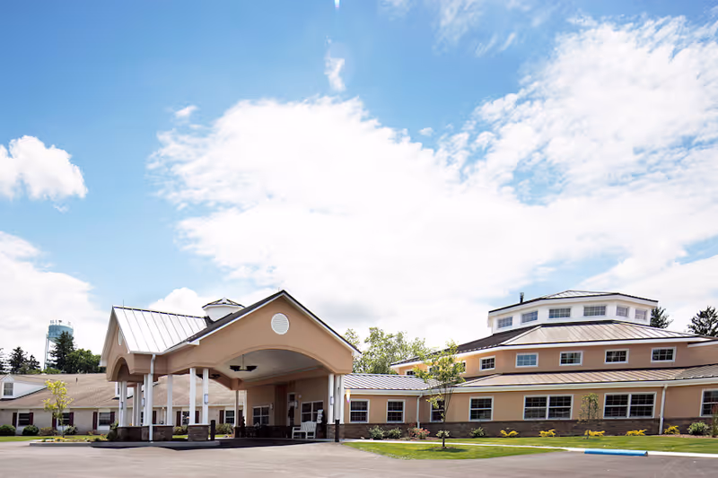 Exterior view of a single-story retirement community building with a covered entrance, beige walls, multiple windows, and a well-maintained lawn under a partly cloudy sky.