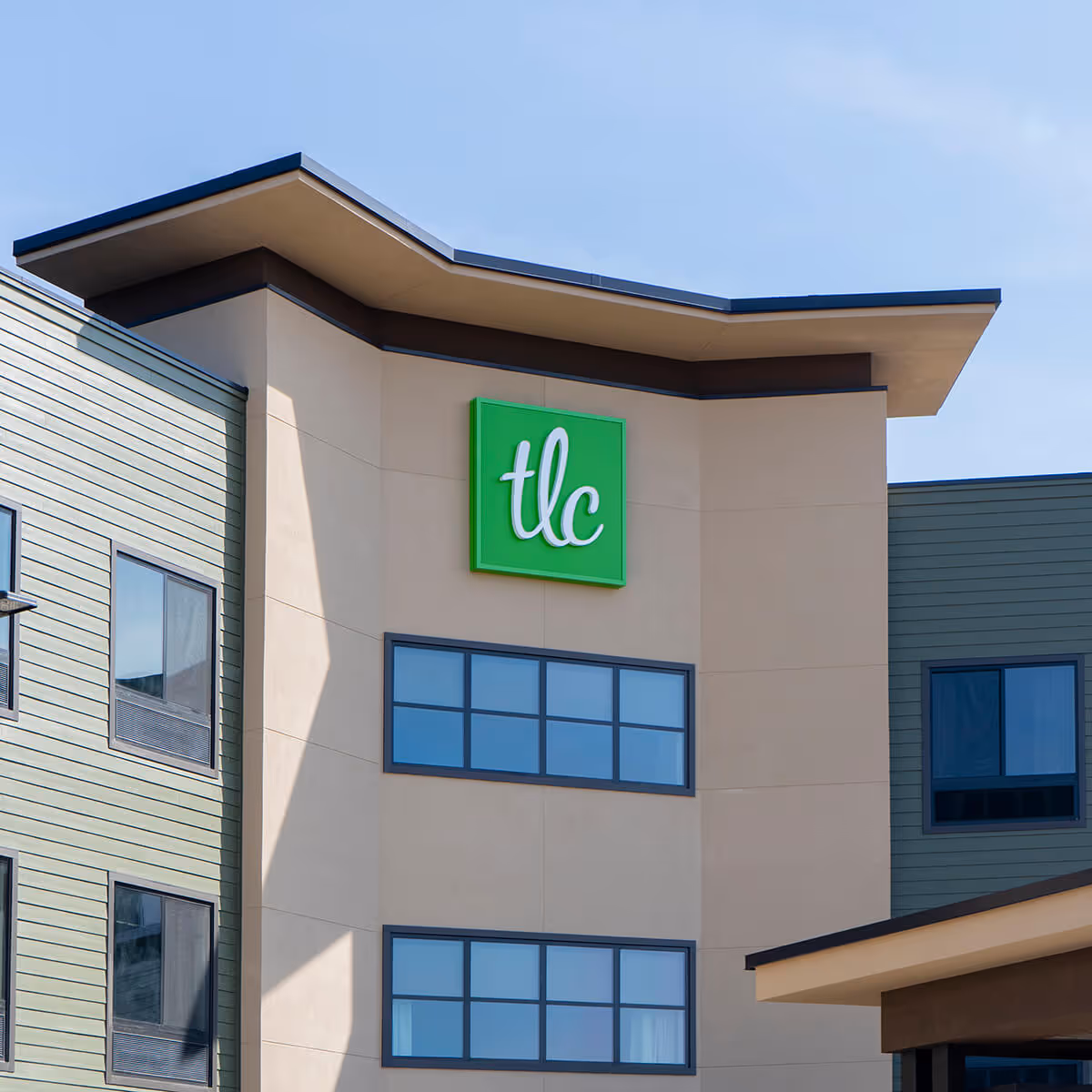 Exterior view of a modern building with beige and green walls, featuring a green sign with white lowercase letters 'tlc' mounted near the top. The building has multiple windows and a unique angular roof design under a clear blue sky.