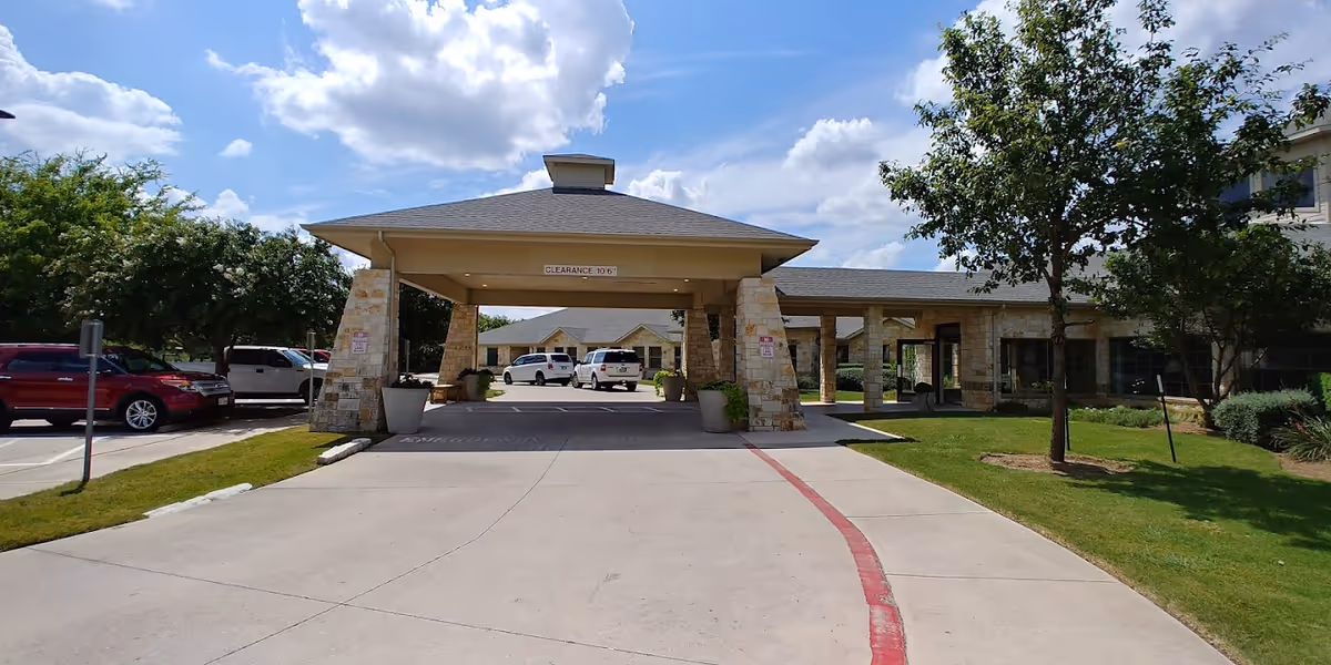 Entrance driveway to Onion Creek Nursing and Rehabilitation Center with a covered drop-off area supported by stone pillars. Several vehicles are parked nearby, and there are trees and green lawns on either side under a partly cloudy blue sky.