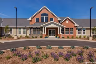 Front exterior view of a single-story senior living facility building with a peaked roof, large windows, and a covered entrance. There is a circular driveway in front with landscaped flower beds containing purple flowers and small shrubs. Two street lamps are visible on either side of the driveway.