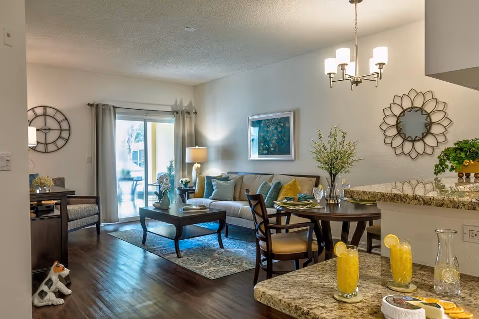 A well-lit living and dining area in a senior living facility featuring a beige sofa with colorful cushions, a wooden coffee table with decorative items, a round dining table set with plates and glasses, a chandelier above, a large wall clock, a decorative mirror, and a sliding glass door leading to an outdoor patio. The room has wooden flooring and granite countertops with two glasses of orange juice and a pitcher.
