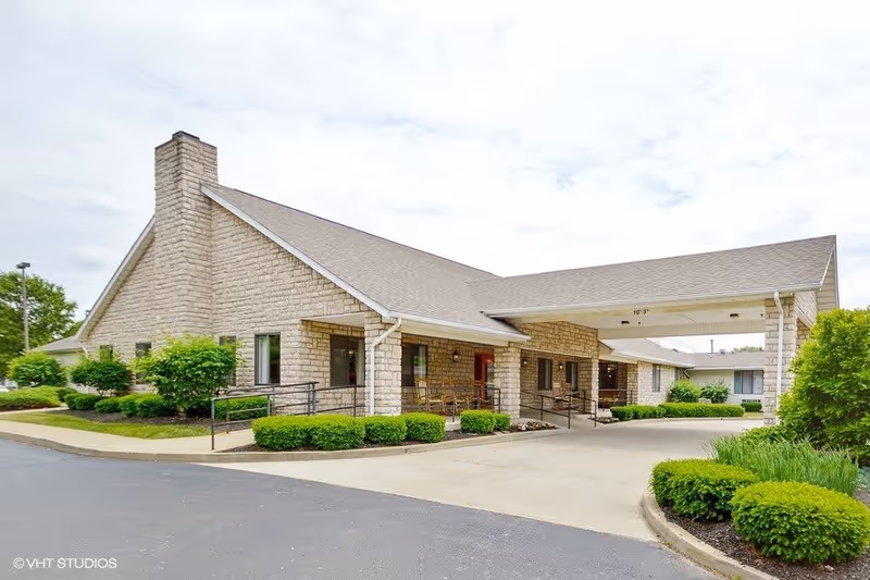 Exterior view of a single-story senior living facility building with stone walls, a sloped roof, and a covered entrance driveway. The entrance area features a ramp and some greenery including bushes and small trees around the building.
