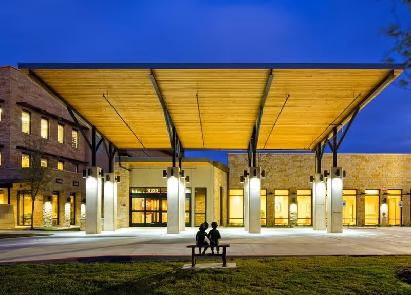 Exterior view of The Watermark at Southpark Meadows building entrance at dusk, featuring a large wooden canopy supported by metal beams and illuminated pillars. Two children sit on a bench in the foreground on a grassy area.