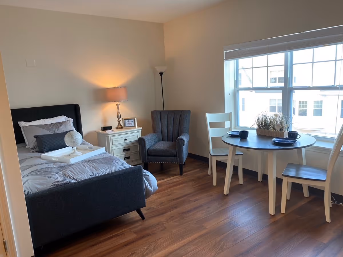 A cozy senior living bedroom with a single bed featuring gray and white bedding, a white nightstand with a lamp and a small framed picture, a gray upholstered armchair, and a round dining table with two chairs near a large window letting in natural light.