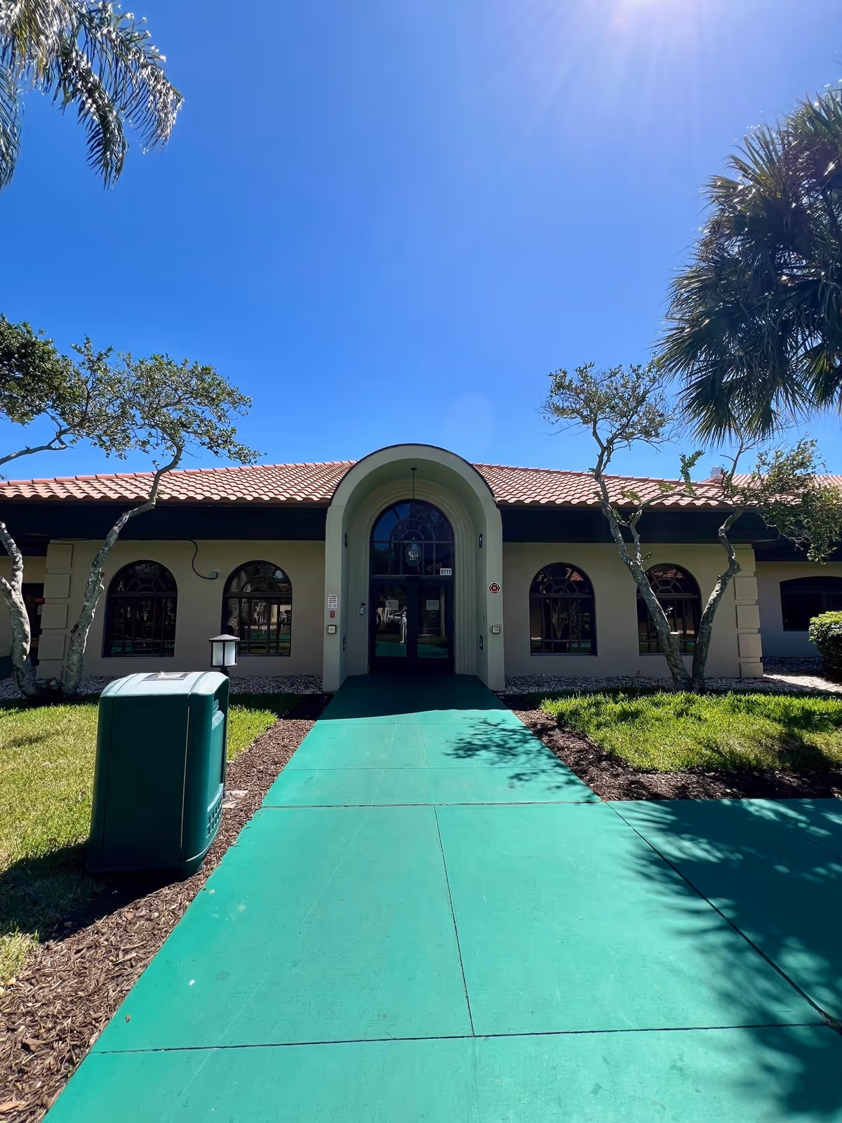 Front exterior view of a single-story building with a red tile roof and arched windows and entrance. A green walkway leads to the entrance, surrounded by grass, small trees, and a green trash bin on the left side. The sky is clear and blue.