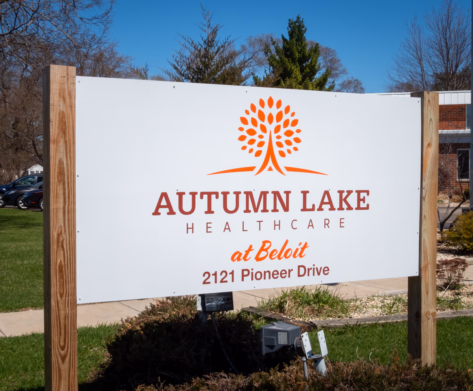 Outdoor sign for Autumn Lake Healthcare at Beloit located at 2121 Pioneer Drive, mounted on wooden posts with a tree logo above the text, with a building and trees in the background under a clear blue sky.
