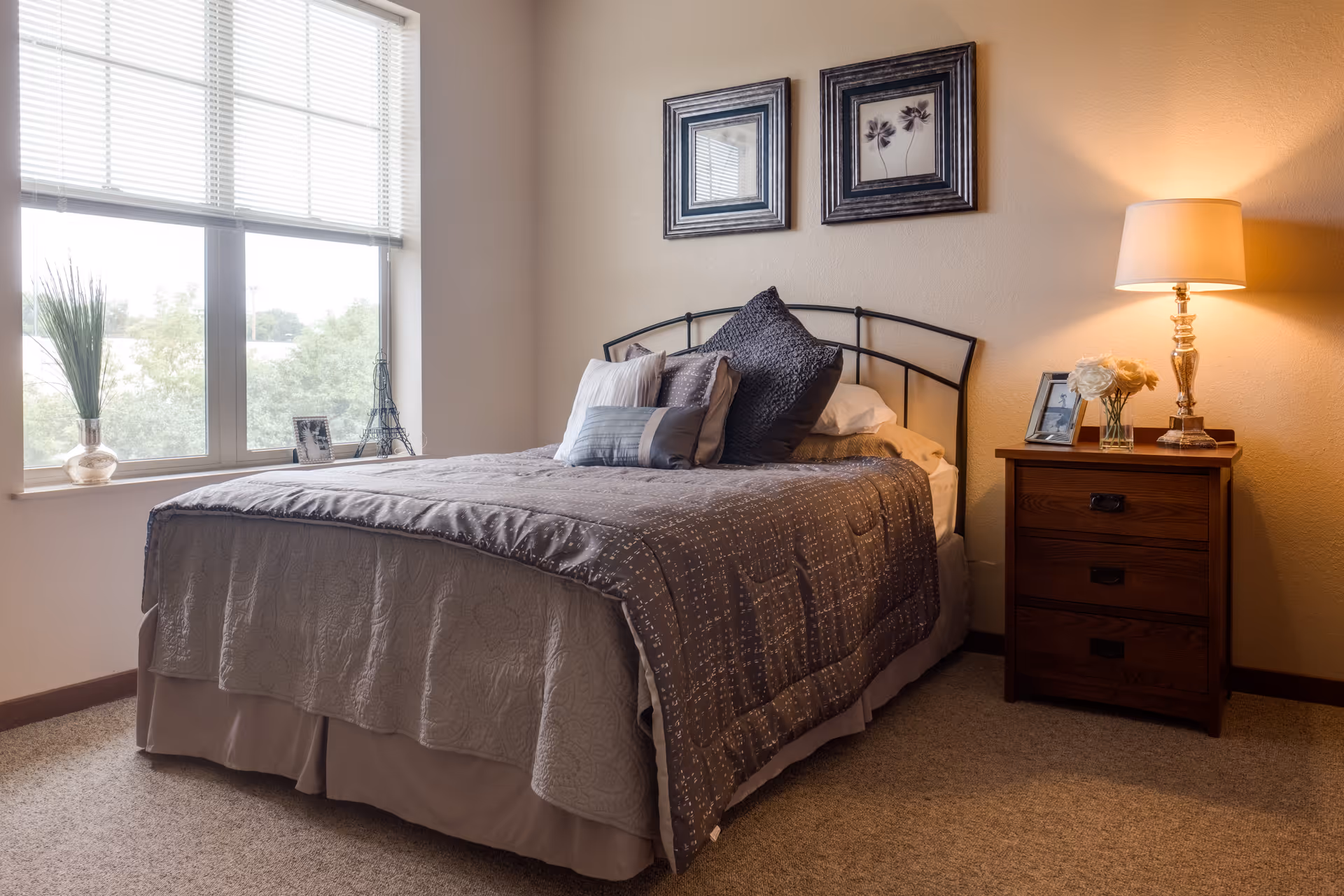 A cozy bedroom with a neatly made bed featuring multiple pillows and a gray quilt. Next to the bed is a wooden nightstand with a lit lamp, a small vase of flowers, and a framed photo. Two framed pictures hang on the wall above the bed. A large window with blinds allows natural light to brighten the room.