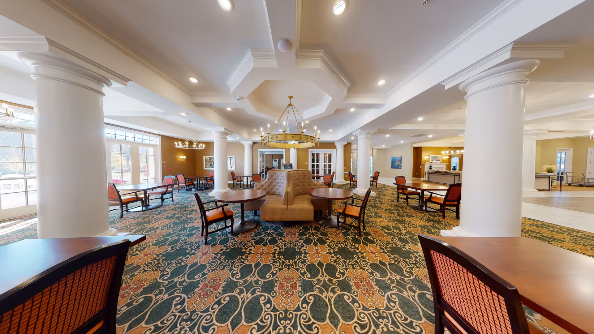 Spacious senior living dining area with patterned carpet, round tables, chairs, and a central tufted booth seating under a large chandelier. The room features white columns, warm lighting, and large windows letting in natural light.