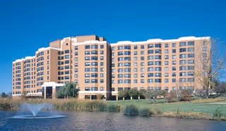 Large multi-story beige building with many windows situated beside a pond with a water fountain, under a clear blue sky.