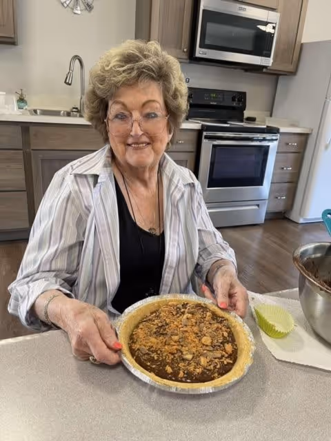 An elderly woman with glasses and curly hair is sitting at a kitchen counter holding a pie topped with nuts and crumbs. Behind her is a modern kitchen with wooden cabinets, a stainless steel oven, microwave, and a sink.