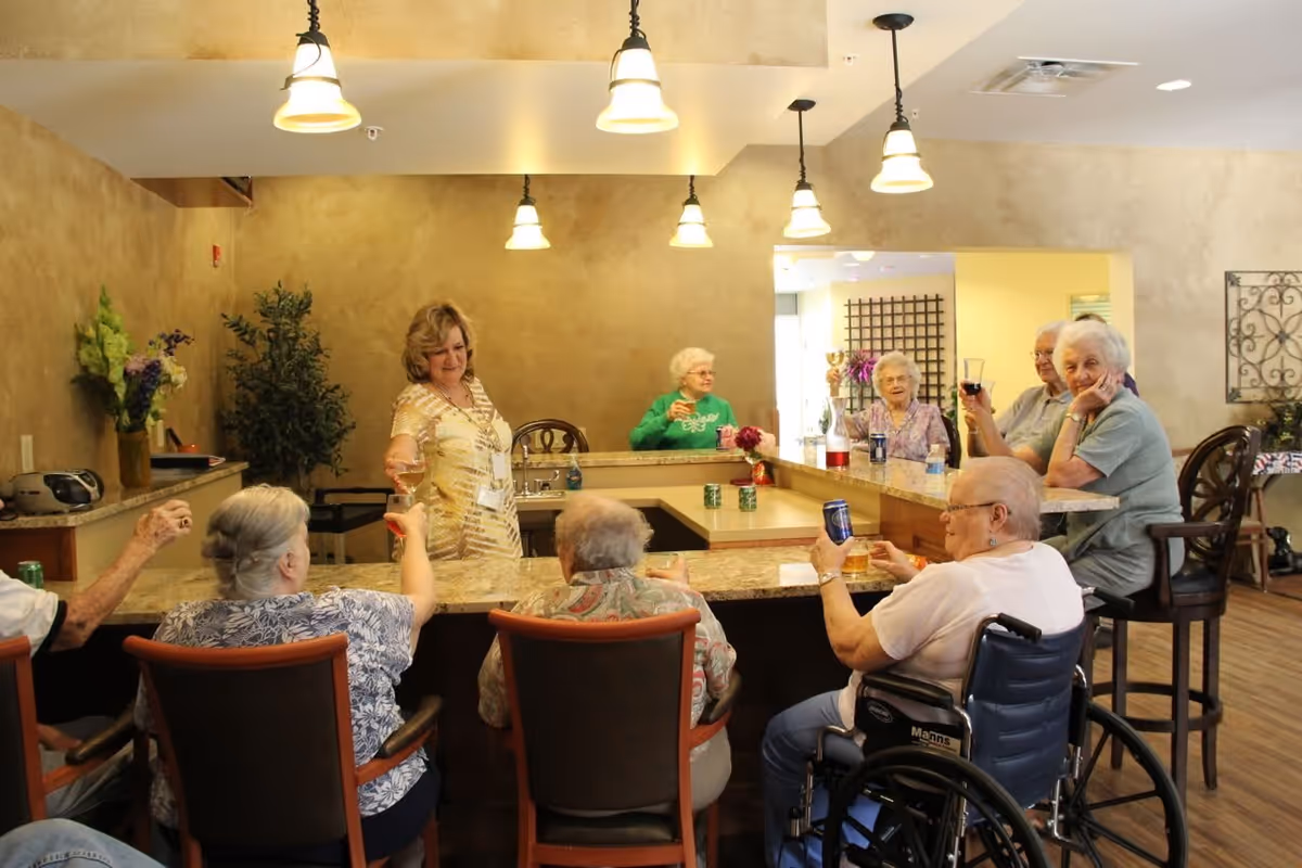 A group of elderly people sitting around a U-shaped bar counter in a well-lit room, raising their glasses and cans in a toast. A woman stands behind the counter serving drinks. The room has warm beige walls, hanging pendant lights, and some plants and flowers as decoration.