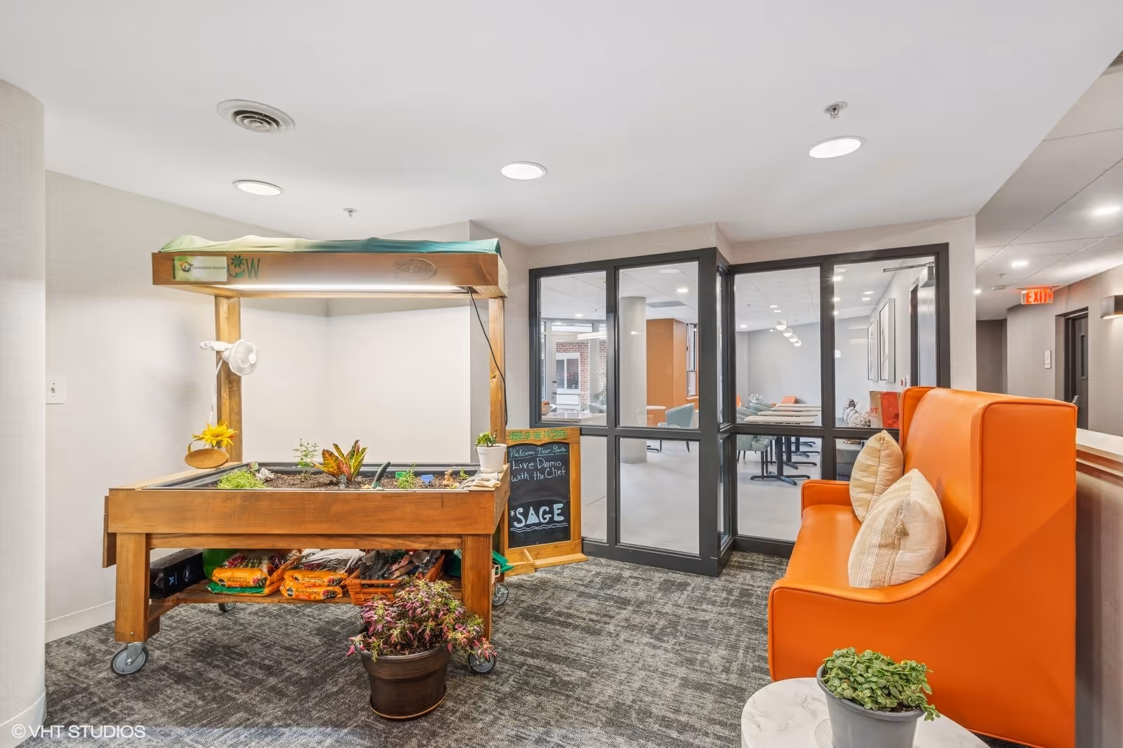 Indoor common area with a wooden planter box on wheels containing plants and gardening supplies. Next to it is a small chalkboard sign advertising a live demo with the chef. There is an orange high-back bench with two pillows on the right side and a small round table with a potted plant in front of it. In the background, glass doors lead to another room with tables and chairs.